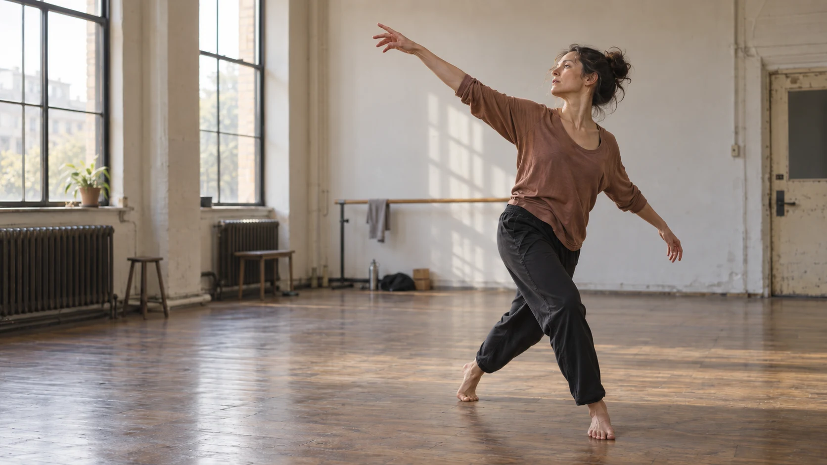 A dancer reaches across a sunlit rehearsal studio.